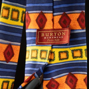 Peut inclure: Cravate en soie à motifs avec un fond bleu, des losanges orange et des carrés jaunes. La cravate porte l'étiquette "BURTON MENSWEAR Pure Silk" sur un fond rouge.