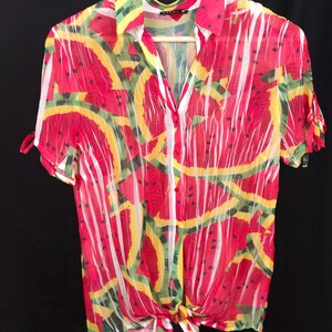 May include: A short-sleeved button-up shirt with a watermelon slice pattern. The shirt features red, yellow, green, and white colors. The shirt is tied at the waist and hangs on a wooden hanger.