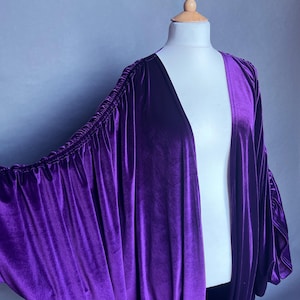 Könnte beinhalten: Ein leuchtend lila Samt-Kimono-Stil-Umhang mit langen, gerafften Ärmeln. Der Umhang ist über einer weißen Schaufensterpuppe drapiert, um sein fließendes Design zu zeigen. Der Text "Eden Jones Co" ist unten sichtbar.