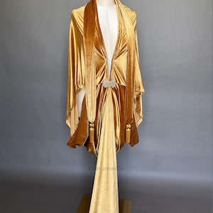 May include: A gold velvet cape with a deep V-neckline, long draped panels, and a central rhinestone brooch closure. The garment features two long hanging fabric strips with tassel ends. The text EdenJonesCo is visible at the top and bottom.