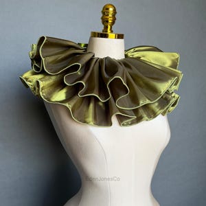 May include: A ruffled, olive green collar with a gold-colored top. The collar is made of a shiny fabric and is displayed on a mannequin. The text "EdenJonesCo" is visible in the image.