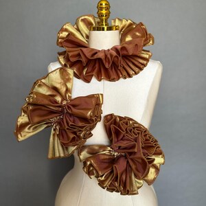 Puede incluir: Un conjunto de tres accesorios con volantes en color dorado metálico y marrón sobre un maniquí. El set incluye un cuello circular y dos piezas a juego con forma de lazo. La tela presenta un acabado brillante y reflectante.