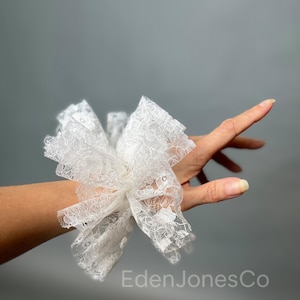 Könnte beinhalten: Ein weißes Spitzen-Armband mit floralem Design. Das Armband besteht aus zarter, geschichteter Spitze und ist für das Tragen am Handgelenk konzipiert. Das Bild ist vor einem grauen Hintergrund aufgenommen.