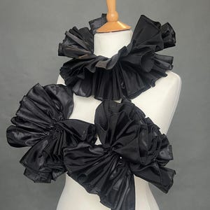 May include: Black ruffled collar and shoulder pieces on a white mannequin. The collar is a large, layered design, and the shoulder pieces are large, ruffled bows. The items are made of a shiny, satin-like material. The text "EdenJonesCo" is visible.