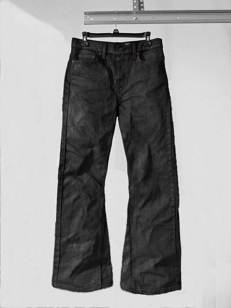 May include: Black denim jeans with a slight flare at the bottom, hanging from a black hanger. The jeans have a classic five-pocket design and a dark, textured appearance. The pants are displayed against a plain white background.