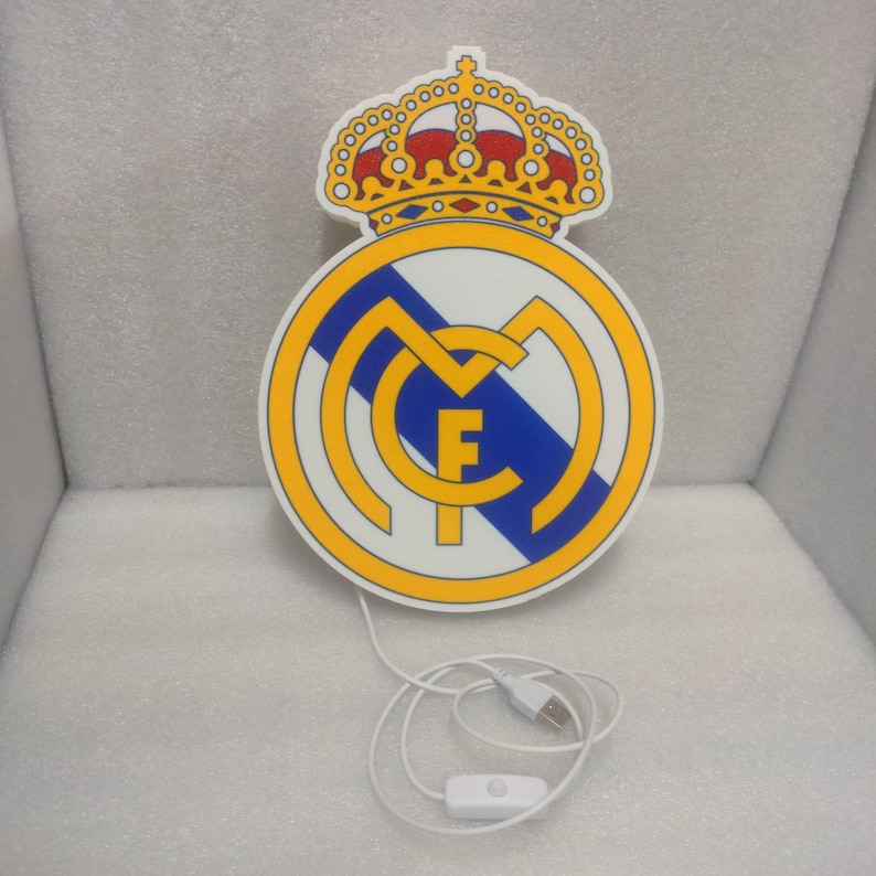 K&ouml;nnte beinhalten: Beleuchtetes Schild mit dem Wappen von Real Madrid in Wei&szlig;, Gelb, Blau und Rot. Das Wappen zeigt eine Krone &uuml;ber einem kreisf&ouml;rmigen Emblem mit den Buchstaben "MCF" und einem blauen Streifen. Mit USB-Kabel und Ein-/Ausschalter.