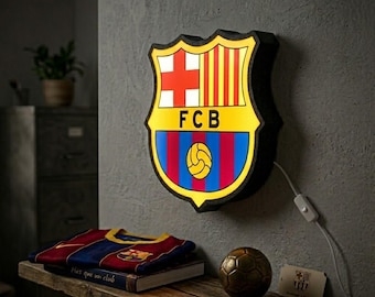 May include: A wall-mounted, illuminated FC Barcelona crest sign featuring the iconic shield shape, red and yellow stripes, a red cross, and a central soccer ball graphic. The sign emits a warm yellow glow and includes a white power cord with a switch.