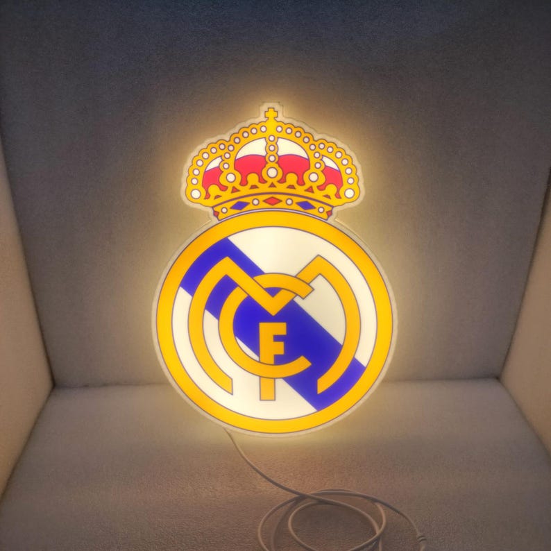 K&ouml;nnte beinhalten: Beleuchtetes Real Madrid Logo mit goldener Krone und rundem Emblem mit blauen und gelben Akzenten. Das Logo ist von innen beleuchtet und erzeugt eine helle Anzeige. Ein wei&szlig;es Kabel erstreckt sich von unten.