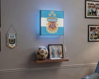 May include: A square, illuminated wall sign featuring the light blue and white Argentine flag with the AFA crest and three gold stars. Below, a wooden shelf holds a soccer ball with the AFA logo and a framed white and light blue soccer jersey.