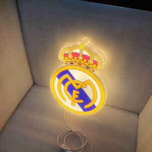 K&ouml;nnte beinhalten: Beleuchtetes Real Madrid-Logo mit goldener Krone, wei&szlig;-blauem Emblem und den Buchstaben "RMF" in einem gelben Kreis. Das Licht wird von einem wei&szlig;en Kabel gespeist.