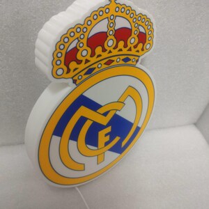 K&ouml;nnte beinhalten: Ein wei&szlig;es, beleuchtetes Schild mit dem Wappen von Real Madrid. Das Wappen zeigt eine goldene Krone mit roten Akzenten &uuml;ber einem kreisf&ouml;rmigen Emblem mit blauem Streifen, goldenem Rand und den Buchstaben "MCF".