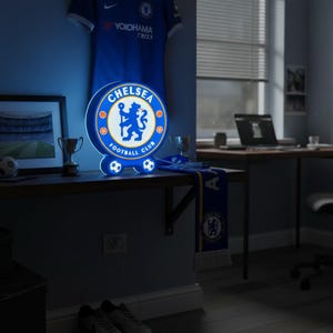May include: A blue and white Chelsea Football Club illuminated sign. The sign features the team's logo and is surrounded by a blue border. The sign is on a shelf with a jersey, trophies, and a scarf.