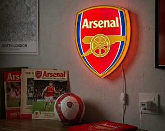 May include: A wall-mounted, illuminated Arsenal Football Club crest sign featuring the team's red, blue, and gold shield logo. The sign glows with a bright red light and is connected to a white power cord plugged into a wall outlet below it.