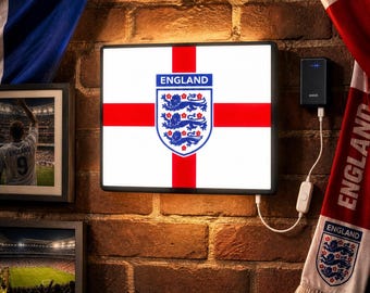 May include: A rectangular light box displaying the England flag and the Three Lions crest with the text ENGLAND. The unit is powered by a white cable connected to a black wall adapter. The design features a white background with a red cross.