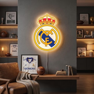 K&ouml;nnte beinhalten: Beleuchtetes Real Madrid-Wappen mit gelbem Umriss, Krone und Teamlogo. Das Wappen ist an einer grauen Wand angebracht. Ein gerahmtes Trikot mit Siemens-Logo und ein Fu&szlig;ball befinden sich darunter.