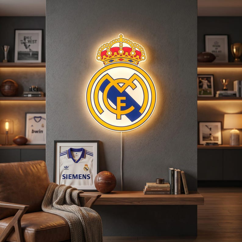 K&ouml;nnte beinhalten: Beleuchtetes Real Madrid-Wappen mit gelbem Umriss, Krone und Teamlogo. Das Wappen ist an einer grauen Wand angebracht. Ein gerahmtes Trikot mit Siemens-Logo und ein Fu&szlig;ball befinden sich darunter.