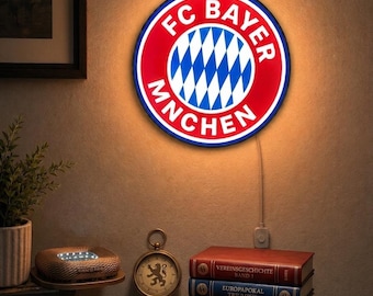 May include: A circular wall-mounted LED sign featuring the FC Bayern München logo in red, white, and blue. The emblem displays the club name in white text around a central diamond pattern. The sign emits a warm glow against the wall surface.