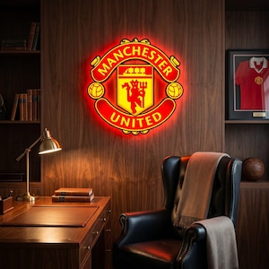 Può includere: Un emblema luminoso del Manchester United, rosso e giallo, è montato su una parete rivestita in legno. La stanza comprende anche una maglia incorniciata, una poltrona in pelle e una scrivania con una lampada in ottone.