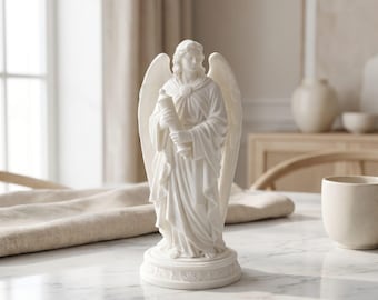 May include: A white angel figurine with outstretched wings, holding a scroll. The statue has detailed facial features and flowing robes. The base is round with a decorative pattern. The angel is standing on a marble surface.