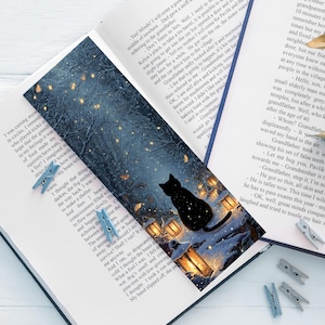 Può includere: Un segnalibro con una scena notturna della foresta, una silhouette di gatto nero e lanterne luminose. Il segnalibro è in un libro aperto con mollette blu.