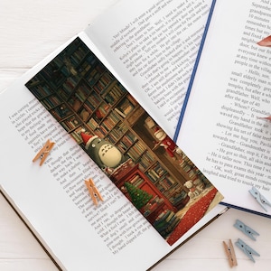 Op de afbeelding: Een open boek met bedrukte tekstpagina's bevat een bladwijzer met een bibliotheekscène en een Totoro-figuur met een rode muts. Verschillende kleine houten wasknijpers liggen verspreid over de pagina's en rechts ligt een takje met roodbruine bladeren.