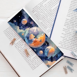 Può includere: Un segnalibro rettangolare con un'illustrazione spaziale di una persona seduta all'interno di una bolla planetaria arancione brillante, circondata da pianeti più piccoli e stelle su uno sfondo blu scuro, posizionato in un libro aperto.