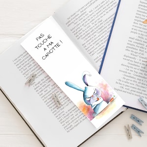 Peut inclure: Un marque-page avec une illustration à l'aquarelle d'un lapin bleu en colère tenant une carotte, avec le texte "PAS TOUCHE A MA CAROTTE!". Le marque-page est placé dans un livre ouvert.