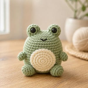 May include: A sage green crocheted frog plush toy with a round cream-colored belly, two black plastic eyes, and a small stitched mouth. The text "no-sew frog" is visible in the upper left corner of the image, written in a dark green font.