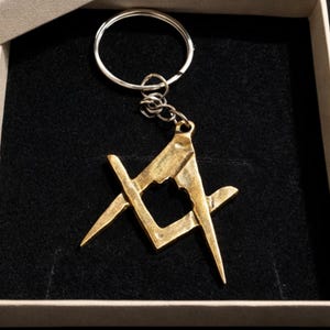 Könnte beinhalten: Ein goldfarbener Metall-Schlüsselanhänger mit einem Freimaurer-Symbol, einem Zirkel- und Winkel-Design. Der Schlüsselanhänger ist an einem silberfarbenen Ring und einer Kette befestigt. Der Artikel wird in einer mit schwarzem Samt ausgekleideten Box präsentiert.