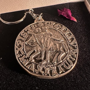 Könnte beinhalten: Ein runder Metall-Schlüsselanhänger mit einem Reliefdesign von zwei Figuren auf einem Pferd, inklusive eines Schildes mit Kreuz. Der äußere Rand ist mit dem lateinischen Text SIGILLUM MILITUM XPISTI versehen und hat einen silberfarbenen Ring.