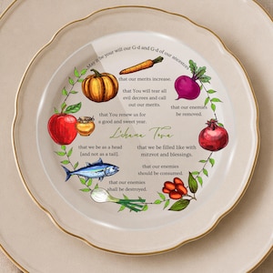 May include: A round ceramic plate with a scalloped gold rim featuring a circular illustration of Rosh Hashanah foods including a pumpkin, carrot, beet, apple, honey, fish, leek, and pomegranate, with English text and the phrase L'chana Tova.