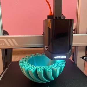 May include: A 3D printer in operation, creating a teal-colored, geometrically patterned bowl. The printer head, labeled "ANYCUBIC", is positioned above the object. The printer is on a black surface.