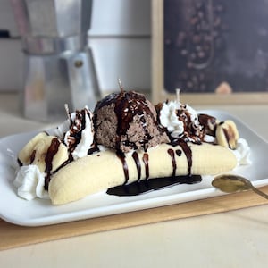 Könnte beinhalten: Eine dekorative Kerze in Form eines Banana Splits. Die Kerze hat eine Bananenbasis, eine Kugel Schokoladeneis, Schlagsahne und Schokoladensauce. Sie wird auf einem weißen rechteckigen Teller präsentiert.