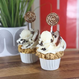 Könnte beinhalten: Zwei Cupcakes mit weißem Zuckerguss, Schokoladenraspeln und einem dekorativen Lutscher. Die Cupcakes stehen in weißen Papierförmchen auf einem Holzbrett. Ein Blumentopf und roter Hintergrund sind sichtbar.