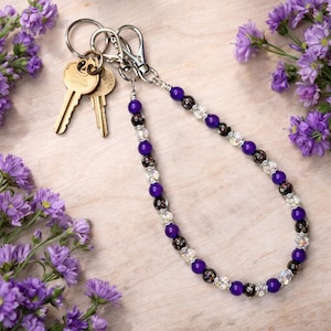 May include: A beaded keychain with a silver clasp and key rings. The keychain features a strand of purple, black, and clear beads. Two gold keys are attached to the key rings. The background is a light wood surface with purple flowers.