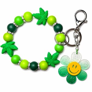 May include: A circular beaded keychain bracelet featuring alternating lime green and dark green silicone beads, interspersed with silver-toned rhinestone spacers and three green leaf-shaped charms. A silver metal clasp holds a glittery flower charm.