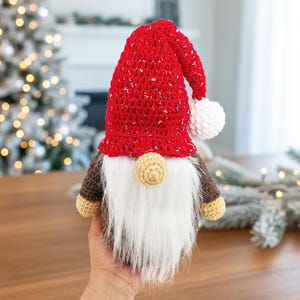 Può includere: Una bambola di gnomo natalizio fatta a mano con un cappello rosso maculato, barba bianca e naso beige. Lo gnomo ha un corpo marrone e braccia e piedi beige. La bambola è tenuta in mano da una persona. Un albero di Natale è sullo sfondo.