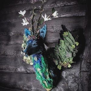 Puede incluir: Una cabeza de ciervo decorativa con tonos azul oscuro y verde. La cabeza de ciervo está adornada con flores blancas y vegetación, creando una exhibición caprichosa y artística. El soporte está fijado a una pared de madera oscura.