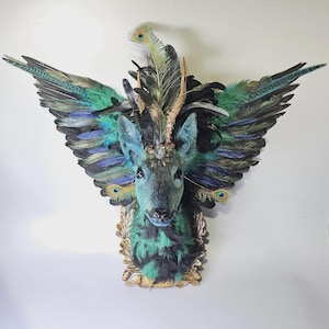 Puede incluir: Una cabeza de ciervo decorativa con plumas verde y turquesa formando alas. La cabeza del ciervo es de color turquesa apagado, con una corona enjoyada. Las plumas incluyen plumas de pavo real. La cabeza está montada sobre una placa dorada ornamentada.