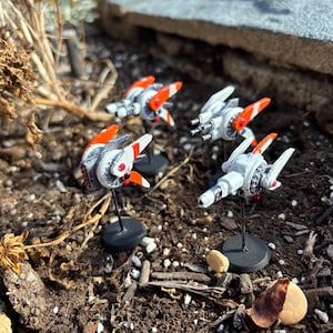 Pode incluir: Quatro modelos em miniatura de naves espaciais futuristas. Cada modelo é branco com detalhes em laranja e vermelho, montado em um suporte preto. Os modelos estão colocados sobre um leito de terra e pequenas pedras, com plantas secas ao fundo.