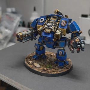 May include: A detailed miniature model of a blue and gold armored robot with a cannon arm and a claw arm. The model is standing on a textured base with small skulls and vegetation. The robot has intricate details and is painted with metallic accents.