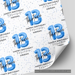 May include: Wrapping paper featuring a repeating pattern of blue balloon-style number 13s, the word thirteen in black script, and the text Happy Birthday To A Special Grandson KIERAN on a white background with small light blue stars.