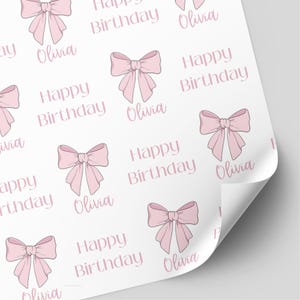 May include: White wrapping paper featuring a repeating pattern of pink bows, the text "Happy Birthday," and the name "Olivia" in a soft pink font. The paper is partially rolled.
