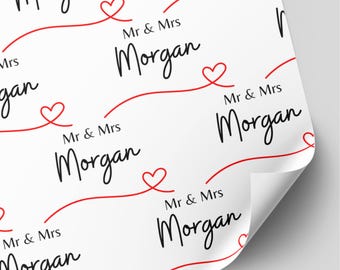 May include: White wrapping paper featuring a repeating pattern of the black text Mr & Mrs Morgan in a script font. The design includes a continuous red line that loops into small heart shapes between each instance of the printed text.