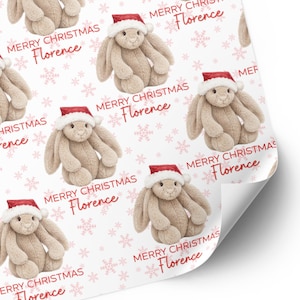 May include: Wrapping paper featuring a repeating pattern of beige plush bunnies wearing red and white Santa hats, interspersed with light pink snowflakes and the red text Merry Christmas Florence printed in a script and sans-serif font combination.