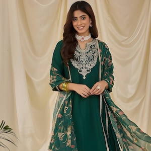 May include: Emerald green salwar kameez ensemble. The tunic features intricate silver embroidery around the neckline and cuffs. The matching pants and dupatta have floral accents. The outfit is paired with gold-toned jewelry and embellished flats.