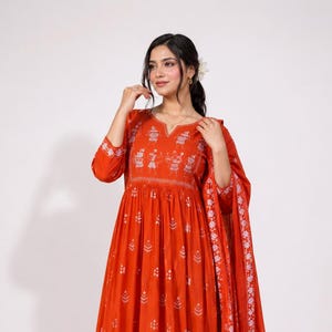 May include: A vibrant orange Anarkali-style dress featuring a notched neckline and three-quarter sleeves. The fabric is adorned with white block-printed patterns, including small floral motifs and traditional figures across the chest and hem.