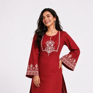 May include: A deep red long-sleeved tunic and matching wide-leg trousers set. The tunic features a round neckline with a small keyhole opening and white floral embroidery on the chest, sleeve cuffs, and hem. The trousers have a scalloped hem.