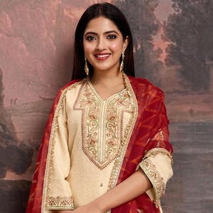 May include: Cream-colored embroidered salwar kameez suit with a red patterned dupatta. The suit features gold embroidery on the neckline, sleeves, and hem. The dupatta has a gold border. The outfit is paired with cream-colored pants and clear heels.