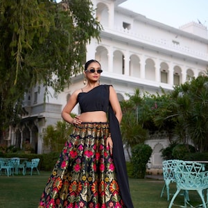 May include: A black crop top and a long skirt with a floral pattern. The skirt features a black background with gold, red, and pink floral designs. The outfit is worn outdoors in front of a white building.